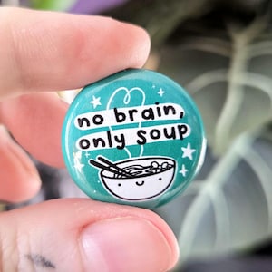 Può includere: Un bottone turchese con la scritta "no brain, only soup" in bianco. Il bottone presenta una ciotola di zuppa a fumetti con bacchette e stelle. Il bottone è tenuto tra due dita.
