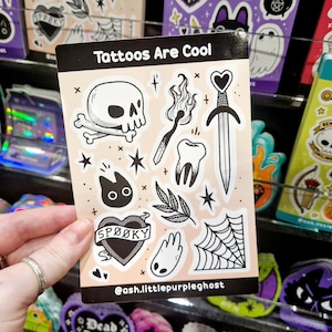 Può includere: Un foglio di adesivi con il testo "Tattoos Are Cool" con varie illustrazioni in bianco e nero. I disegni includono un teschio, un fiammifero, un dente, una spada, un gatto, un cuore con il testo "Spooky", una foglia, un fantasma e una ragnatela.