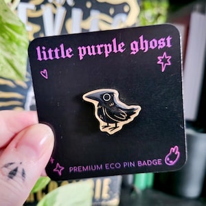 May include: A black enamel crow pin with gold detailing, displayed on a black card. The card features the text "little purple ghost" in purple, along with other text and symbols. The pin is a premium eco pin badge.