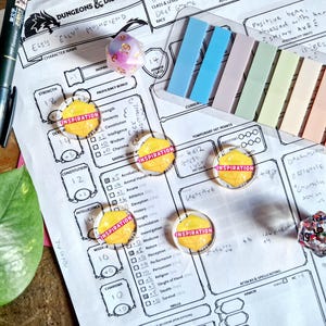 May include: A tabletop role-playing game setup featuring a character sheet, dice, and markers. The sheet has character stats and the word "Inspiration" on several tokens. Colorful sticky notes and a pen are also present.