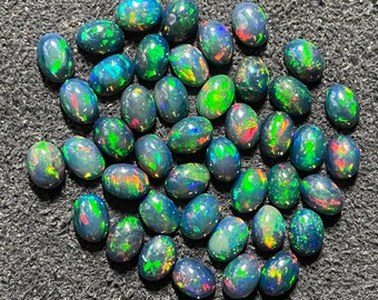 Puede incluir: Una colección de ópalos negros de forma ovalada con destellos iridiscentes verdes, azules y rojos. Los ópalos están dispuestos en un montón suelto sobre una superficie gris oscura.