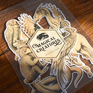 Puede incluir: Un paquete de pegatinas con varias criaturas míticas, como un búho, un grifo y una serpiente alada. Las pegatinas son en tonos marrones y beige, con la inscripción "Magical Creatures Sticker Pack".