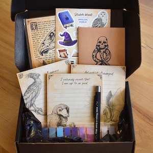 May include: A black cardboard box containing stationery items including lined paper with illustrations of a bird, a platypus-like creature, and a dragon, a sticker sheet with a book and owl, a brown card with a skull, and a black pen with white text.