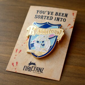 May include: A gold-colored enamel pin featuring a white rabbit wearing a blue scarf and holding a book. The pin is shaped like a shield with the words "Rabbitpaw" written on a banner above the rabbit. The pin is on a brown card with the text "You've been sorted into" and "Firelfanz Design".