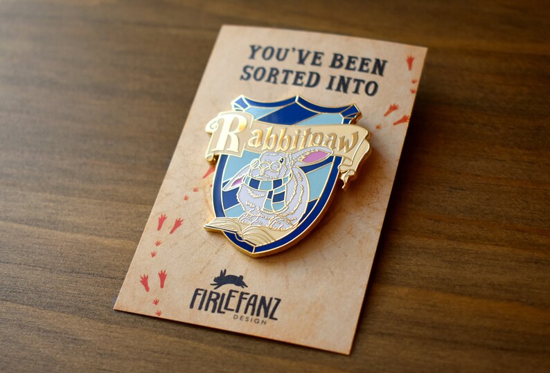 May include: A gold-colored enamel pin featuring a white rabbit wearing a blue scarf and holding a book. The pin is shaped like a shield with the words "Rabbitpaw" written on a banner above the rabbit. The pin is on a brown card with the text "You've been sorted into" and "Firelfanz Design".