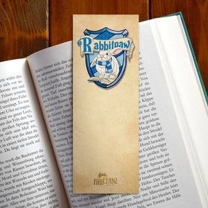 May include: A bookmark featuring a white rabbit wearing glasses and a blue scarf. The rabbit is sitting on a book with a blue and gold shield behind it. The shield has the words "Rabbitpaw" written on it. The bookmark is made of paper and has a brown background.
