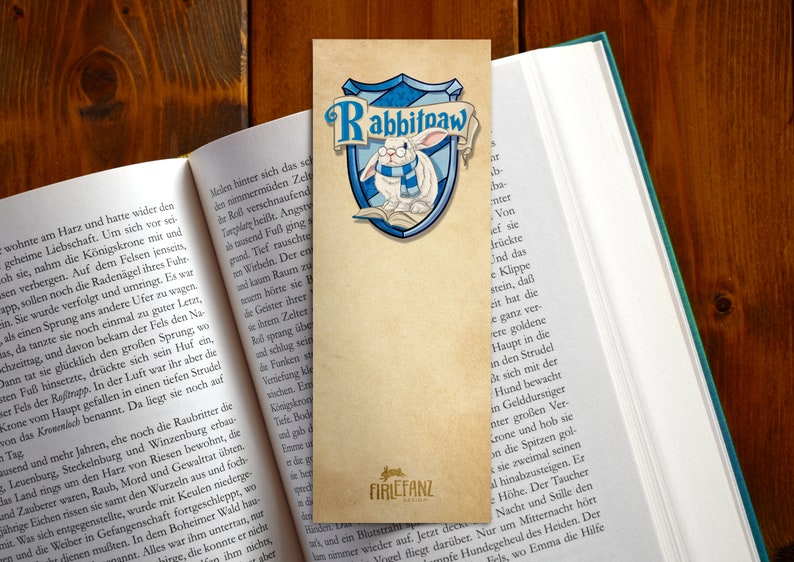 May include: A bookmark featuring a white rabbit wearing glasses and a blue scarf. The rabbit is sitting on a book with a blue and gold shield behind it. The shield has the words "Rabbitpaw" written on it. The bookmark is made of paper and has a brown background.