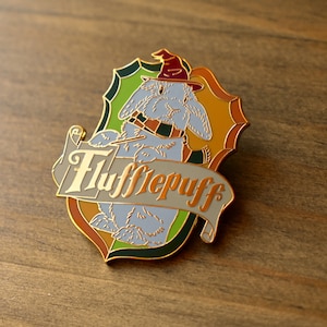 May include: A gold-colored enamel pin featuring a cartoon rabbit wearing a wizard hat and holding a wand. The rabbit is surrounded by a green, yellow, and orange shield with the words "Hufflepuff" written in gold on a white banner.