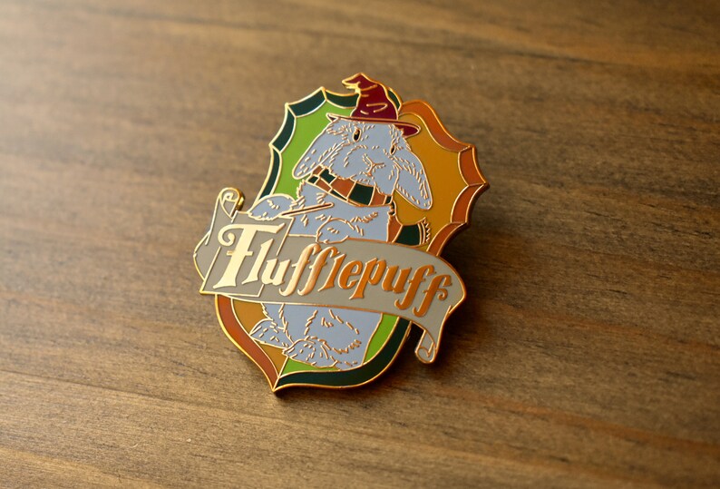 May include: A gold-colored enamel pin featuring a cartoon rabbit wearing a wizard hat and holding a wand. The rabbit is surrounded by a green, yellow, and orange shield with the words "Hufflepuff" written in gold on a white banner.
