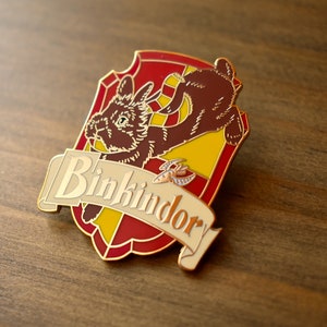 May include: A gold-colored enamel pin featuring a brown rabbit with a yellow and red shield. The shield has the text "Binkindory" on a white banner.