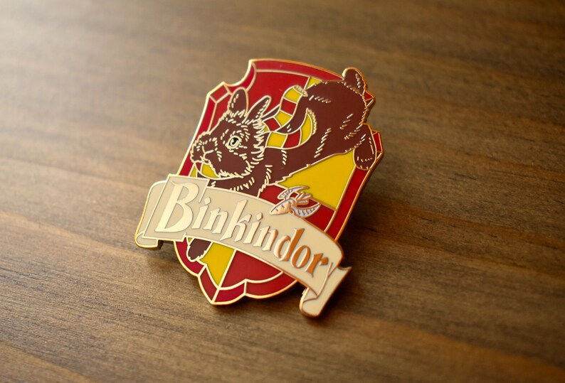 May include: A gold-colored enamel pin featuring a brown rabbit with a yellow and red shield. The shield has the text "Binkindory" on a white banner.