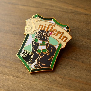 May include: A gold-colored enamel pin featuring a black rabbit wearing a green and white scarf, holding a cauldron with a green liquid. The pin is shaped like a shield with a green and white checkered pattern and the text "Snifferin" in gold lettering.