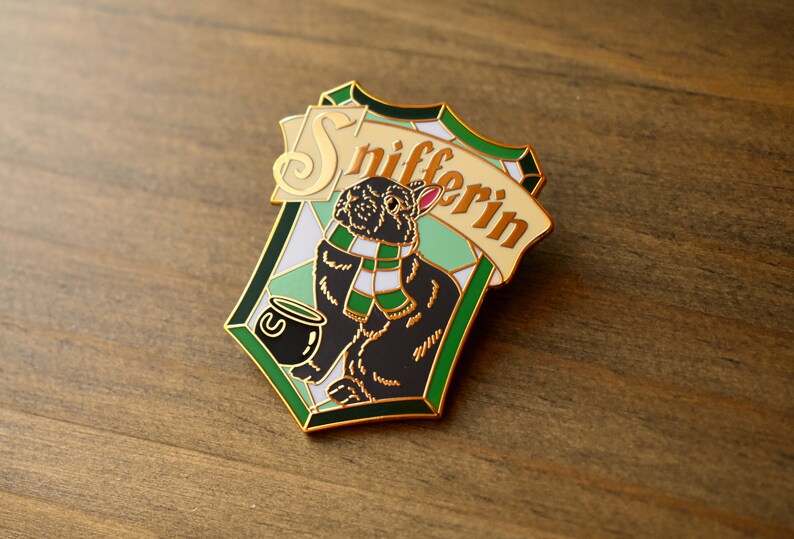 May include: A gold-colored enamel pin featuring a black rabbit wearing a green and white scarf, holding a cauldron with a green liquid. The pin is shaped like a shield with a green and white checkered pattern and the text "Snifferin" in gold lettering.