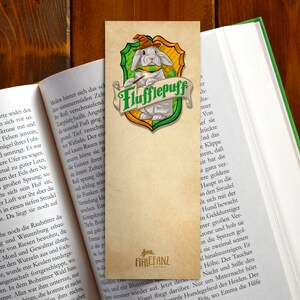 May include: A bookmark featuring a white rabbit wearing a wizard's hat and a green scarf. The rabbit is holding a paintbrush and is sitting on a green and gold shield with the words "Flufflepuff" written on it. The bookmark is made of paper and has a brown background.