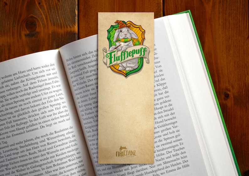 May include: A bookmark featuring a white rabbit wearing a wizard's hat and a green scarf. The rabbit is holding a paintbrush and is sitting on a green and gold shield with the words "Flufflepuff" written on it. The bookmark is made of paper and has a brown background.