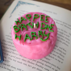 Könnte beinhalten: Eine rosa Kerze mit grünen Buchstaben mit der Aufschrift "HARPEE BIRTH DAE HABBY" liegt auf einem offenen Buch. Eine einzelne weiße Kerze befindet sich in der Mitte der Kerze. Das Buch hat Text auf den Seiten und einen blauen Buchrücken.