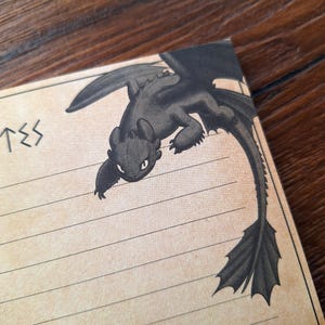 May include: A notepad with lined pages and a black dragon illustration in the upper corner. The dragon has large wings and a long tail. The word "TES" is written in a stylized font on the left side of the notepad.