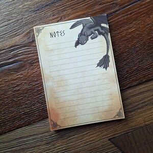 May include: A lined notepad with the word "NOTES" at the top and a black dragon illustration. The notepad has a vintage, aged paper appearance with decorative corner designs. The dragon illustration is black.