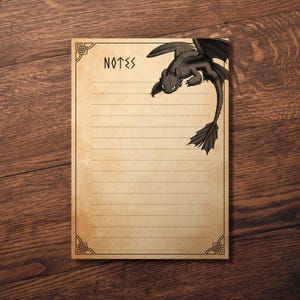Può includere: Un blocco note a righe con la parola "NOTES" in alto e un'illustrazione di un drago nero nell'angolo in alto a destra. Il blocco note ha un bordo decorativo ed è appoggiato su una superficie di legno.