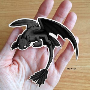 Puede incluir: Una pegatina de drag&oacute;n negro con contornos blancos, con grandes alas y una larga cola. La pegatina mide 10 x 9,4 cm. El drag&oacute;n est&aacute; en posici&oacute;n tumbada, con las alas extendidas. La pegatina se sostiene en una mano.