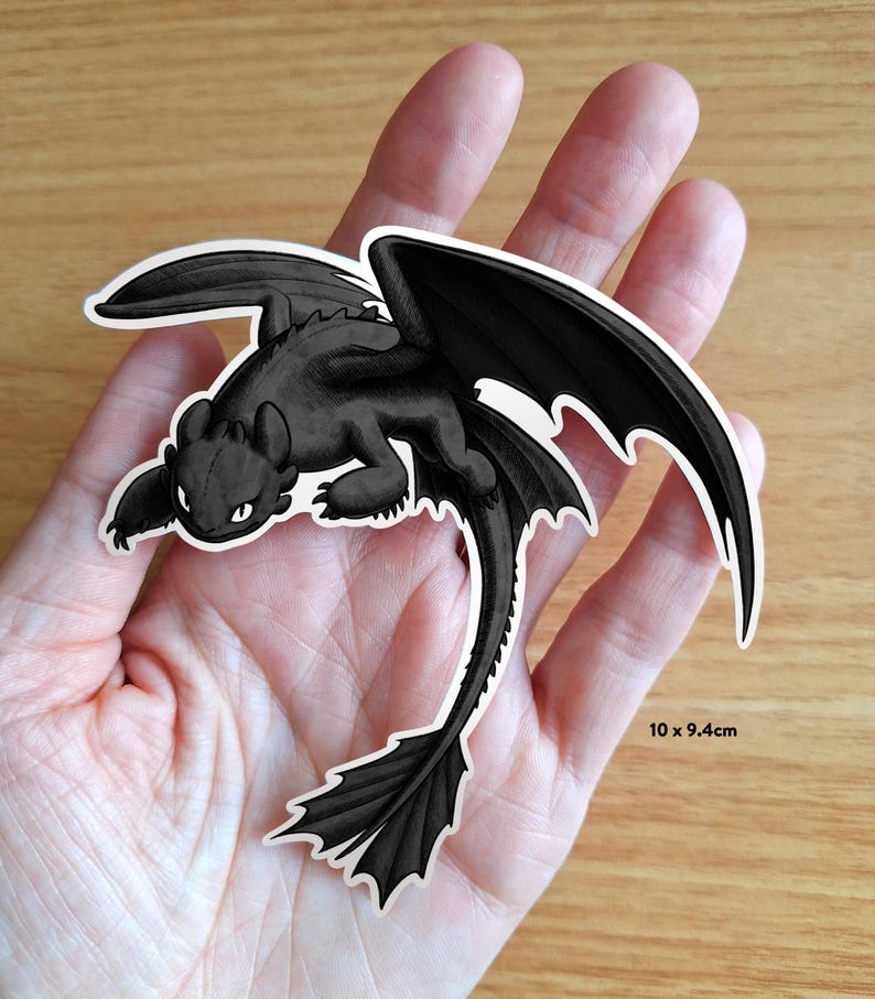 Puede incluir: Una pegatina de drag&oacute;n negro con contornos blancos, con grandes alas y una larga cola. La pegatina mide 10 x 9,4 cm. El drag&oacute;n est&aacute; en posici&oacute;n tumbada, con las alas extendidas. La pegatina se sostiene en una mano.