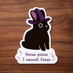Peut inclure: Un autocollant découpé représentant un lapin noir uni portant un chapeau de sorcière violet. Le lapin a un œil et un nez blancs simples. Sous le lapin, le texte Hocus pocus I cannot focus est écrit en lettres violettes avec des étoiles.