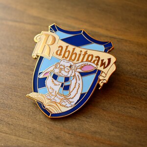 May include: Gold enamel pin featuring a white rabbit wearing glasses and a blue and white scarf, sitting on a book. The pin is shaped like a shield with a blue and white background and the text "Rabbitpaw" in gold lettering.