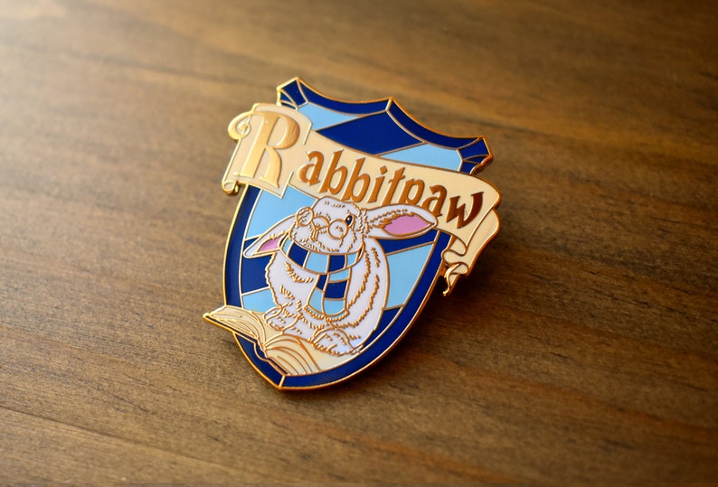 May include: Gold enamel pin featuring a white rabbit wearing glasses and a blue and white scarf, sitting on a book. The pin is shaped like a shield with a blue and white background and the text "Rabbitpaw" in gold lettering.