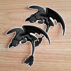 Puede incluir: Dos pegatinas negras de drag&oacute;n con contornos blancos se muestran sobre una superficie de madera marr&oacute;n claro. Los dragones tienen alas grandes, orejas puntiagudas y una cola larga. Las pegatinas est&aacute;n en una pose de vuelo.