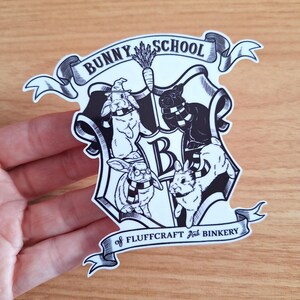 May include: Black and white sticker of a bunny school crest. The crest features a shield with four bunnies wearing scarves and a banner that reads "Bunny School of Fluffcraft and Binkery".