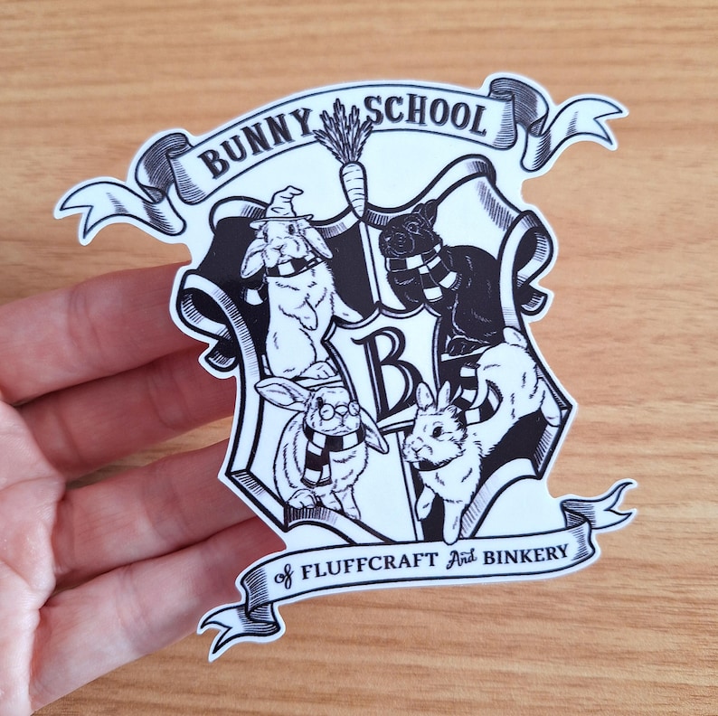 May include: Black and white sticker of a bunny school crest. The crest features a shield with four bunnies wearing scarves and a banner that reads "Bunny School of Fluffcraft and Binkery".
