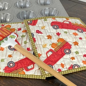May include: A quilted fabric potholder featuring a pattern of red pickup trucks carrying pumpkins and autumn leaves on a white wood-grain background, with a green checkered border, paired with a silicone spatula with a pumpkin print.