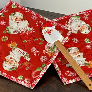 May include: Two quilted fabric pot holders featuring a red background with a repeating pattern of Santa Claus faces, candy canes, and snowflakes. A wooden rolling pin lies beneath the pot holders, and a silicone spatula with a Santa head is placed on top.