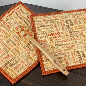 May include: Two square quilted pot holders featuring a cream background with orange and brown text including words like Harvest, Blessings, and Hello Fall, paired with a wooden spatula with a pumpkin-patterned silicone head and Wilton branding.