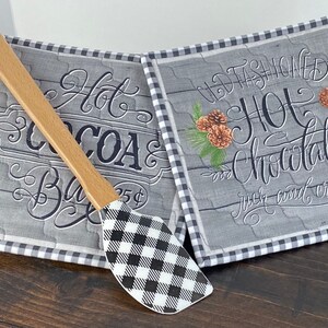 May include: Two square fabric pot holders with a gray wood-grain pattern, black and white gingham trim, and decorative text reading Hot Cocoa Bar 25 cents and Old Fashioned Hot Chocolate rich and creamy, paired with a wooden handled spatula.