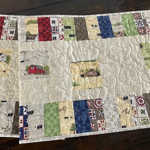 May include: A rectangular quilted table runner featuring a patchwork design with farm-themed fabric squares. The quilt displays illustrations of red barns, silos, and cows, interspersed with geometric star patterns in red, blue, green, and brown tones.