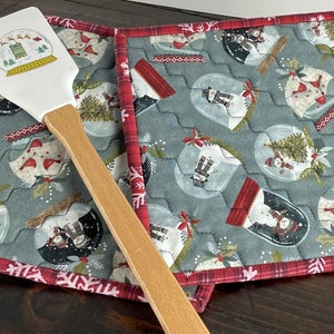 May include: A white silicone spatula with a snow globe graphic featuring Santa and reindeer, resting on two quilted fabric pot holders. The pot holders have a gray background with a repeating snow globe pattern and red plaid trim.