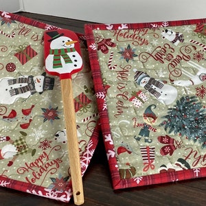 May include: Two square quilted potholders featuring a beige background with Christmas-themed illustrations including snowmen, nutcrackers, gifts, and text like Merry and Bright, alongside a wooden-handled spatula with a snowman graphic.