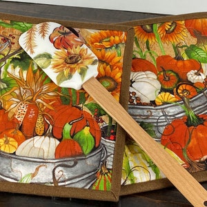 May include: Two square fabric pot holders with brown trim featuring a print of pumpkins, sunflowers, and galvanized metal buckets. A silicone spatula with a matching sunflower print and a wooden handle rests diagonally across the pot holders.