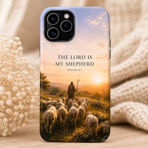 May include: A smartphone case featuring a printed landscape scene of a shepherd walking with a flock of sheep at sunset. The text "THE LORD IS MY SHEPHERD" and "PSALM 23:1" is printed in black lettering above the figure in the center of the case.