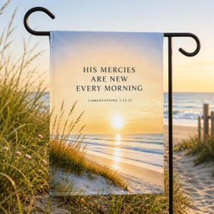 Puede incluir: Una bandera de jardín rectangular colgada en un soporte de metal negro, con un amanecer sobre una playa de arena con hierba de duna. El texto dice: HIS MERCIES ARE NEW EVERY MORNING LAMENTATIONS 3:22-23 en una fuente serif oscura en el cielo.