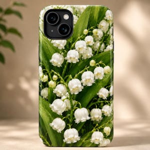 Puede incluir: Una funda rectangular para smartphone con un estampado botánico detallado de flores de lirio de los valles blancas con pequeñas flores en forma de campana y hojas de color verde vibrante. El diseño cubre toda la parte trasera, incluida la cámara.