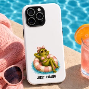May include: A white smartphone case featuring a graphic of a green frog wearing a pink polka dot bikini, a matching bow, and sunglasses while lounging in a pink and white striped pool float, with the text JUST VIBING printed below the illustration.