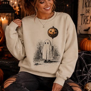 May include: Beige crewneck sweatshirt featuring a Halloween-themed graphic. The graphic depicts a ghost holding a jack-o'-lantern balloon. The design is in black and white, with a detailed illustration of a tree and the ghost's form. The sweatshirt is a neutral color.