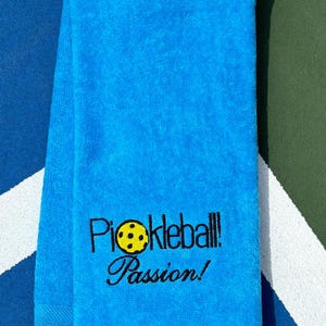 May include: A bright blue towel with the text "Pickleball! Passion!" embroidered in black. A yellow pickleball is embroidered in the text.