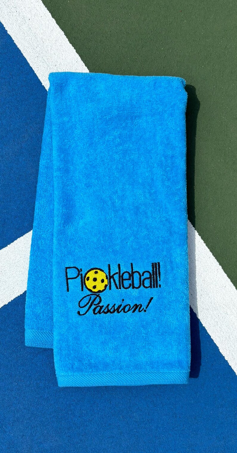 May include: A bright blue towel with the text "Pickleball! Passion!" embroidered in black. A yellow pickleball is embroidered in the text.