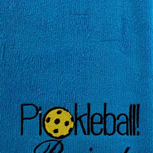 May include: A blue towel with black embroidery that reads "Pickleball! Passion!" and features a yellow pickleball with black stitching.