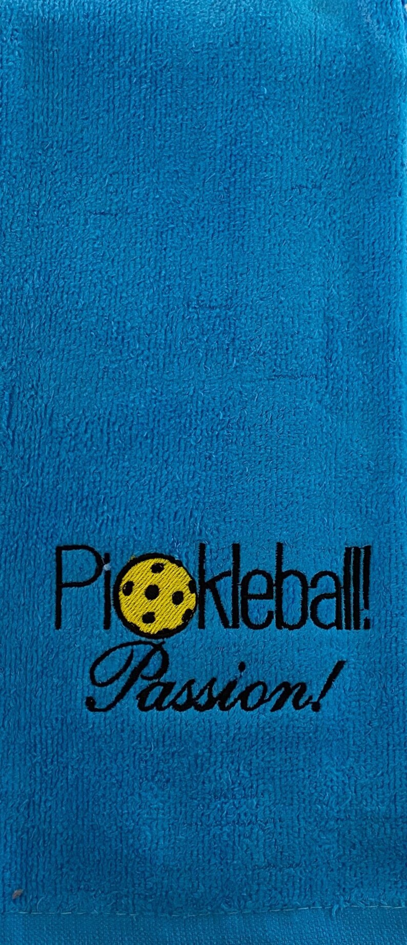 May include: A blue towel with black embroidery that reads "Pickleball! Passion!" and features a yellow pickleball with black stitching.