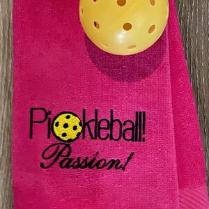 May include: Pink towel with the text "Pickleball Passion!" embroidered in black. A yellow pickleball is on top of the towel.