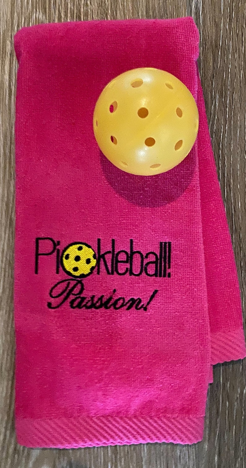 May include: Pink towel with the text "Pickleball Passion!" embroidered in black. A yellow pickleball is on top of the towel.